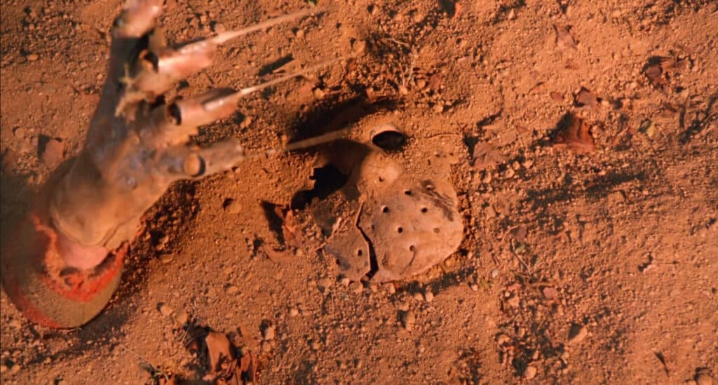 Freddy Krueger's gloved hand grabbing Jason's mask and pulling it in to the dirt.