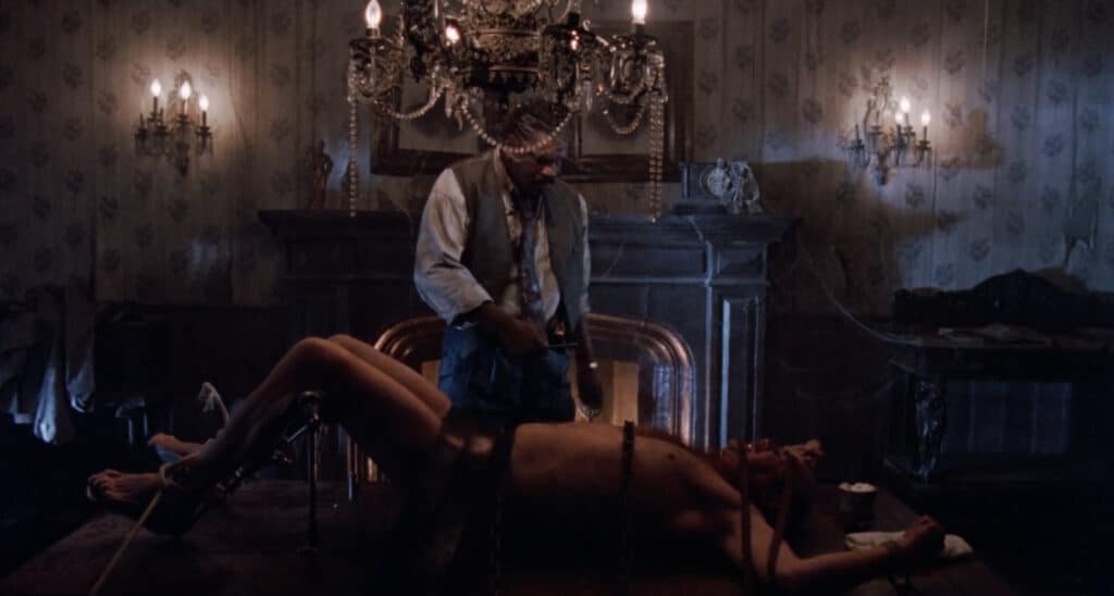 A man tied down on a table while another man stands over him with a knife.